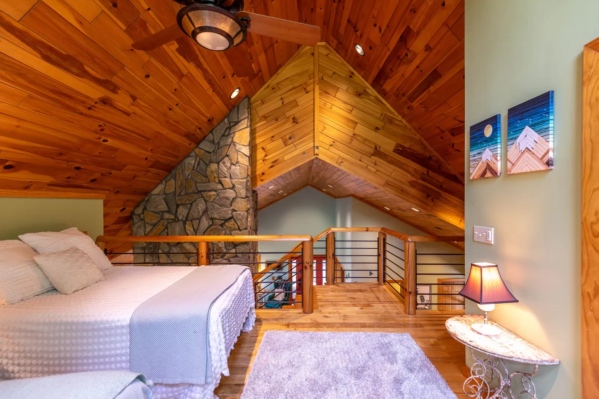 A comfortable lower-level bedroom with a plush bed accented by decorative pillows, a stone wall, a wooden railing and staircase, a ceiling fan for comfort, and a soft glow from a decorative lamp. The space feels restful and thoughtfully designed for relaxation.