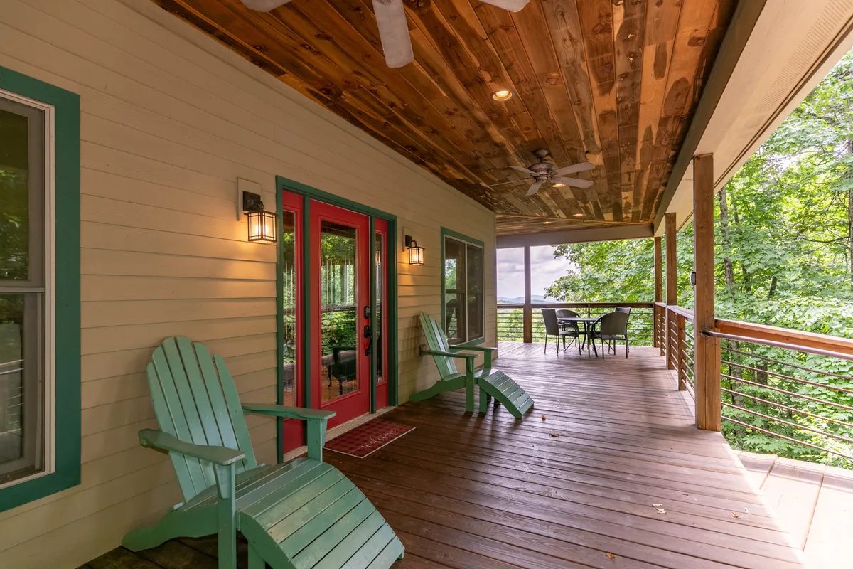 This image captures a cozy outdoor seating area on a deck, featuring inviting Adirondack chairs, a striking bright red door, a wooden ceiling overhead, and lush greenery surrounding the space. The scene feels tranquil and connected to the outdoors, suggesting a perfect spot to enjoy mountain air with your dog.