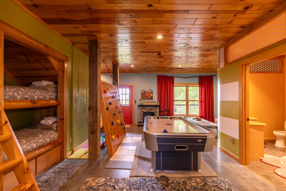 A wide angle of the family zone, this image shows bunk beds, a colorful climbing wall, the air hockey table, and a cozy fireplace. Large windows and red curtains brighten the space, while the fireplace and seating area create a warm, inviting mood for families to gather and relax after play.