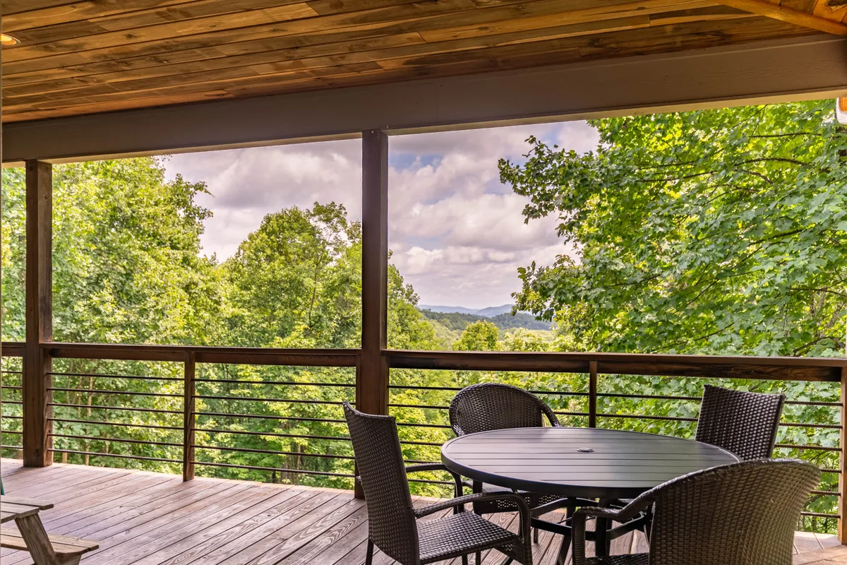 A serene outdoor deck with a circular table and woven chairs, framed by a view of trees and distant mountains. The space invites guests to move seamlessly from the great room to the outdoors, enjoying meals or quiet moments while immersed in natural beauty.