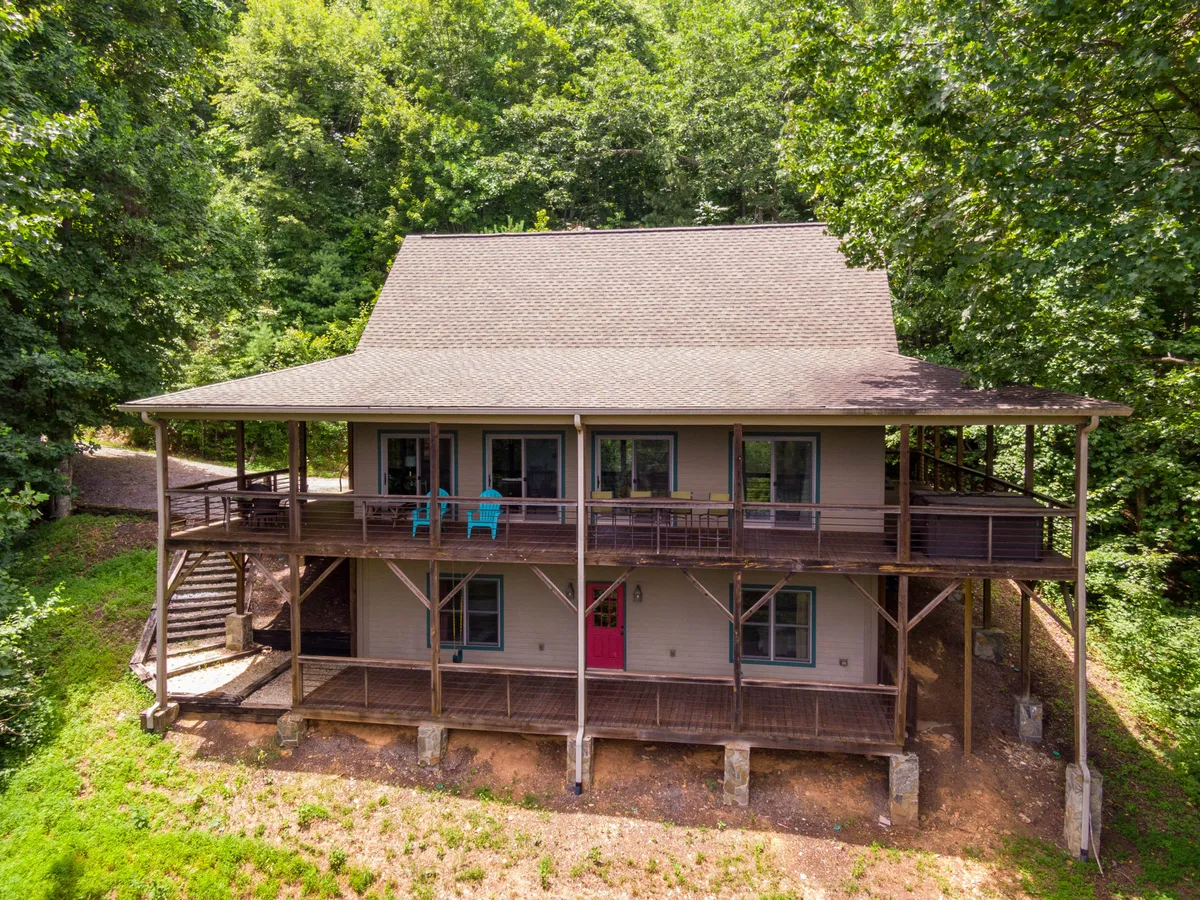 A large, welcoming porch featuring a bright pink door, wooden deck, and blue chairs. This inviting space is surrounded by greenery and perfectly suited for morning coffee, group gatherings, or enjoying the peaceful neighborhood ambiance.