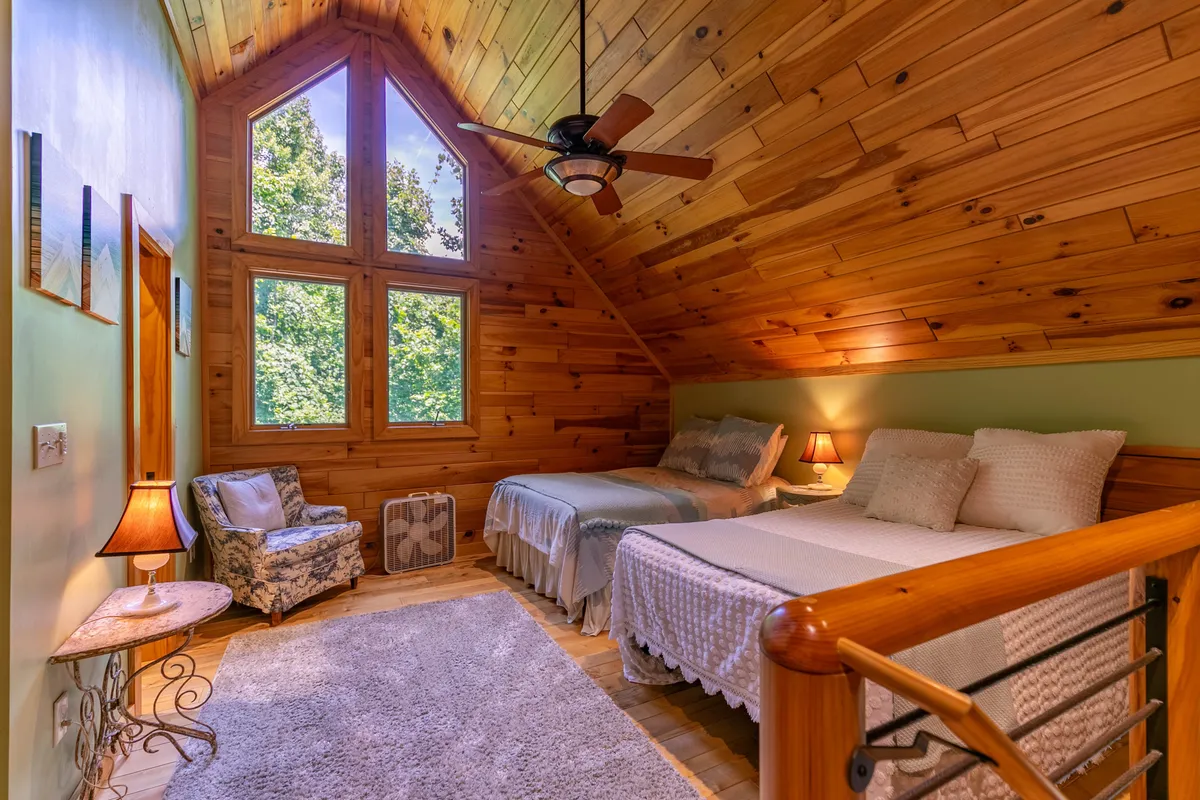A loft-style bedroom featuring two full beds with soft linens, large windows overlooking the trees, plenty of natural wood accents, and a comfortable sitting area. The natural light and calming decor create a perfect retreat for older kids or friends.