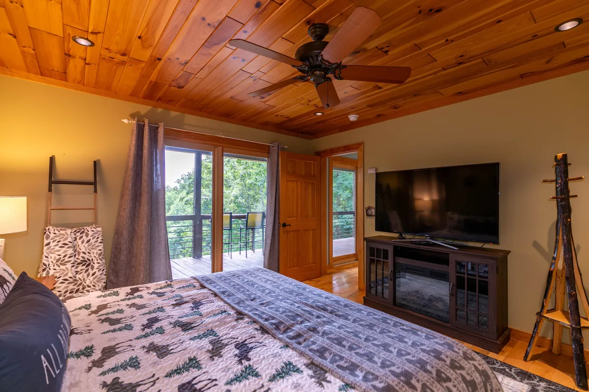 A tranquil bedroom with a wooden ceiling, large windows that frame woodland views, a bed with patterned bedding, and a TV unit. The space feels airy and inviting with plenty of natural light and a cozy ambiance.