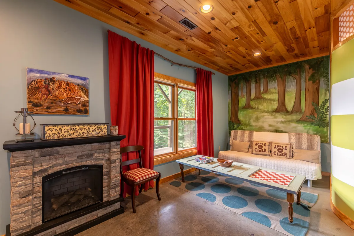 This image offers a view of a fireside seating area with a window that brings in a scenic view, a cozy sofa, a colorful mural, and playful cushions. The room is set up for relaxation, conversation, and enjoying the surrounding natural beauty from indoors.