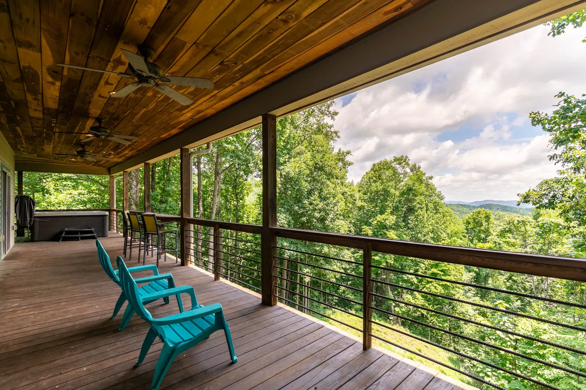 A spacious wooden deck furnished with vibrant blue chairs, surrounded by lush trees and opening up to a scenic mountain view. The deck is positioned for sunset watching and stargazing, offering ample space for groups to gather and connect with nature.