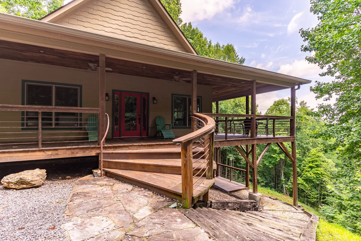 The image presents a spacious wooden deck with comfortable chairs, a curved wooden staircase, red doors, and views of the surrounding natural landscape. The setting is welcoming and serene, ideal for relaxing, socializing, or letting dogs explore under supervision.