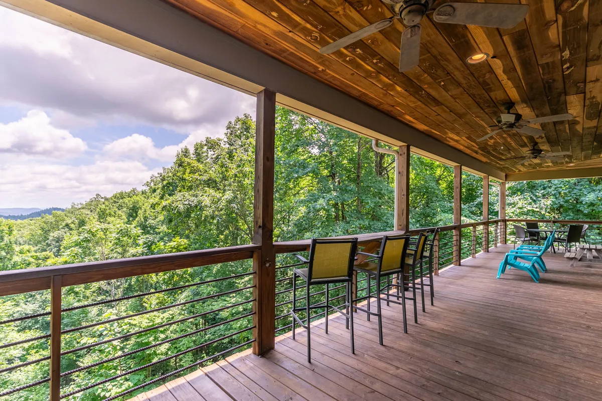 A serene exterior view of the cabin's wooden deck, furnished with bar stools and overlooking a peaceful forested landscape. The open-air setting invites guests to socialize, enjoy sunsets, or simply take in the beauty of the outdoors.