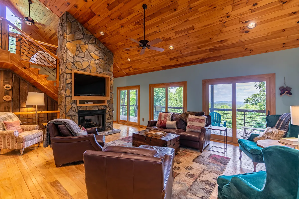 A spacious great room bathed in natural light, this image features a striking stone fireplace as its centerpiece. Plush leather seating is arranged for conversation and relaxation, while large windows offer breathtaking views of the surrounding mountains. The vaulted wooden ceiling adds rustic elegance, and a coffee table invites gatherings or board games.