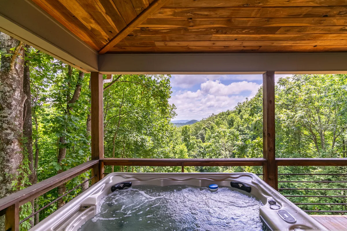 This image captures a private hot tub nestled on a wooden deck, surrounded by lush greenery with sweeping mountain views in the background. The tranquil atmosphere invites relaxation, whether soaking at sunset or under the stars. The deck area is spacious, ideal for socializing or quiet moments with nature.