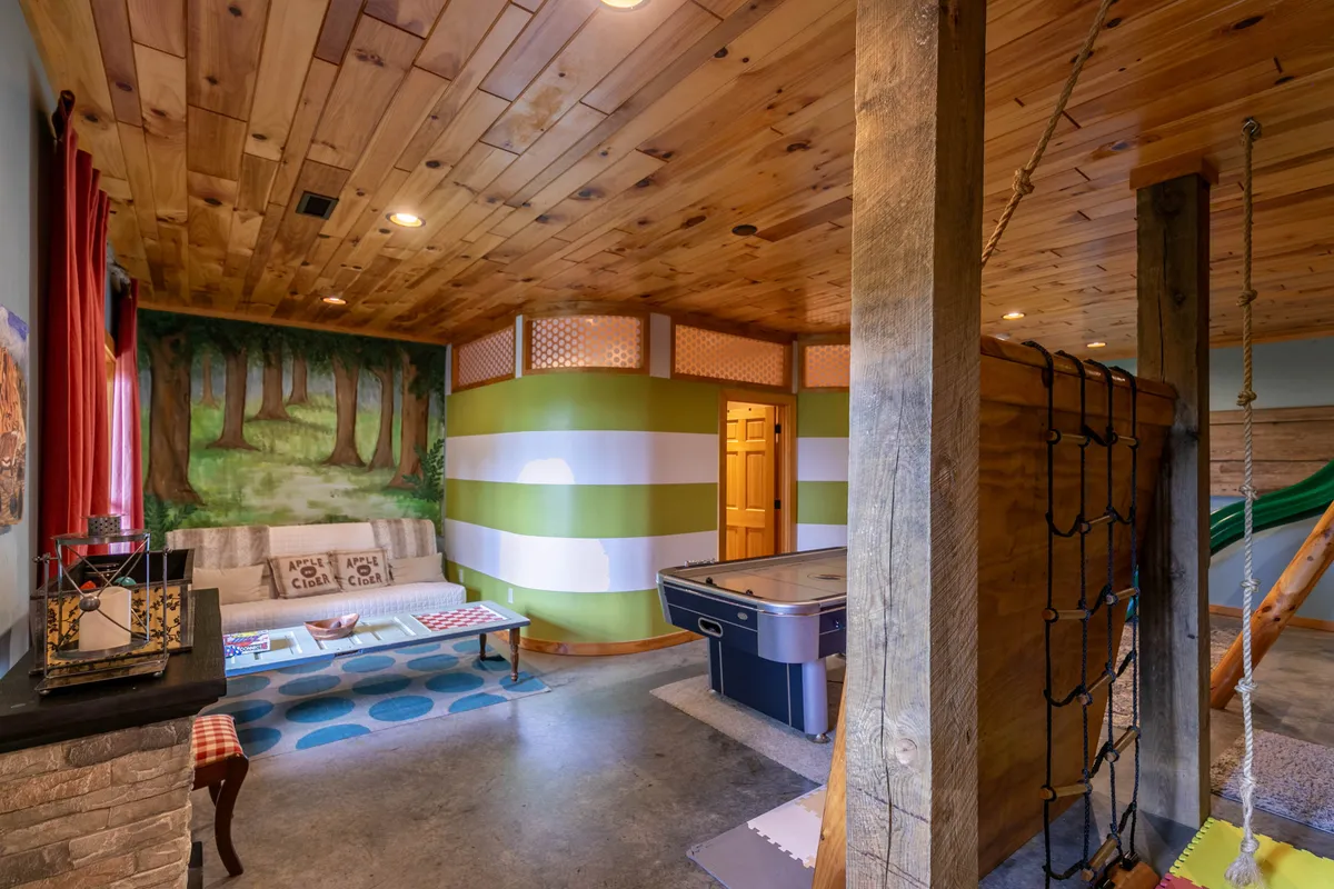 A vibrant indoor family play area featuring a colorful forest mural on the wall, a custom play structure, a hot tub, and a comfortable seating area. The space radiates fun and relaxation for both kids and adults.