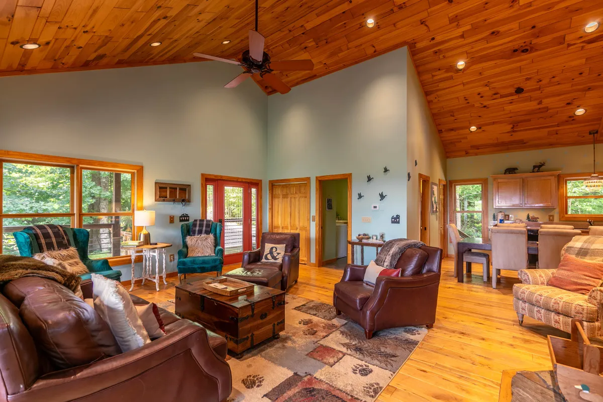This image captures a spacious living area with a wooden ceiling, leather chairs, a central coffee table, large windows, and colorful decor. The room is filled with natural light and arranged for both relaxing and socializing, evoking a sense of warmth and gathering.