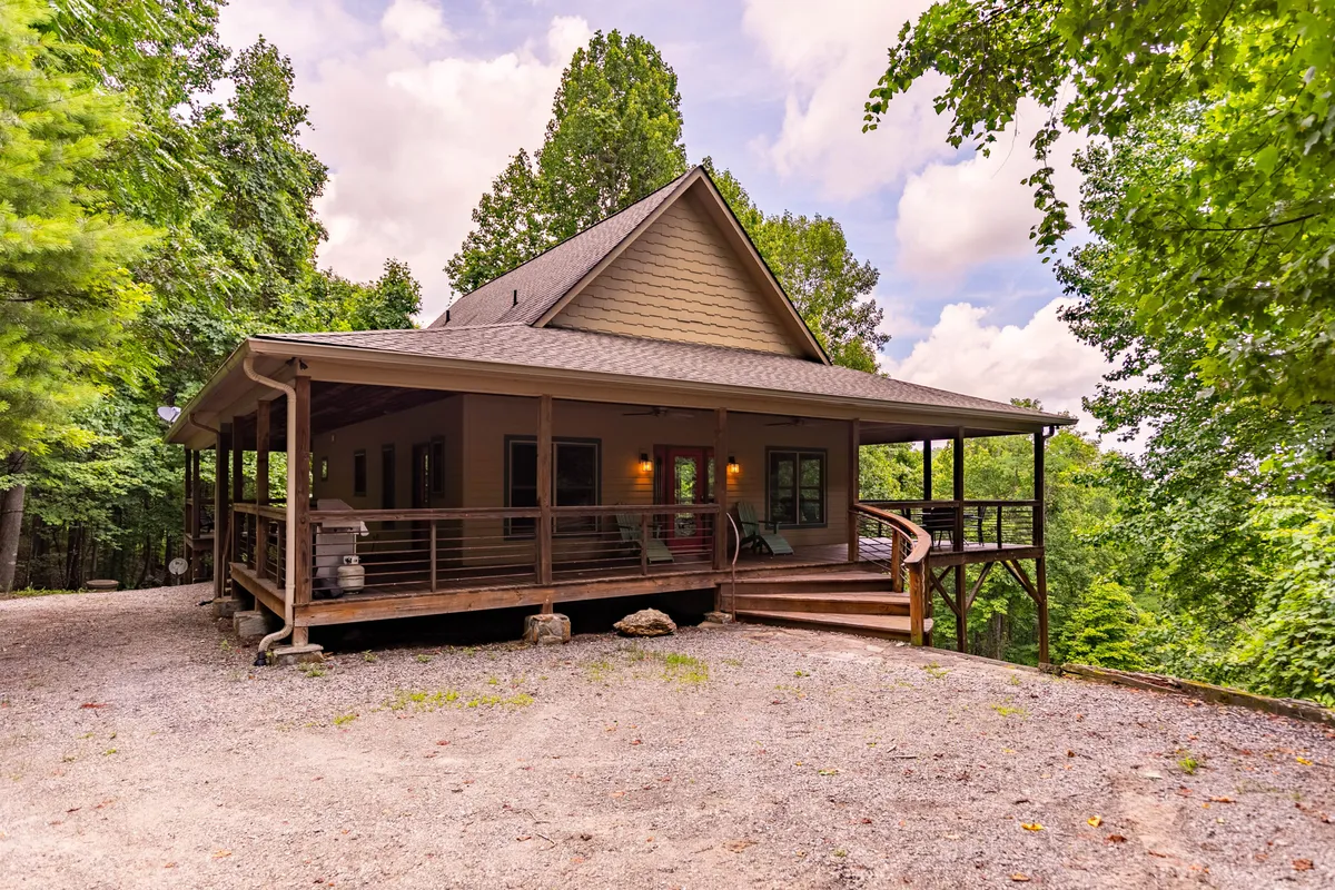 The image showcases the cabin's inviting front porch framed by a distinctive roofline, set against a backdrop of towering trees and tranquil mountain scenery. The porch hints at leisurely evenings spent outdoors, while the lush, private woodland setting promises peace and seclusion.