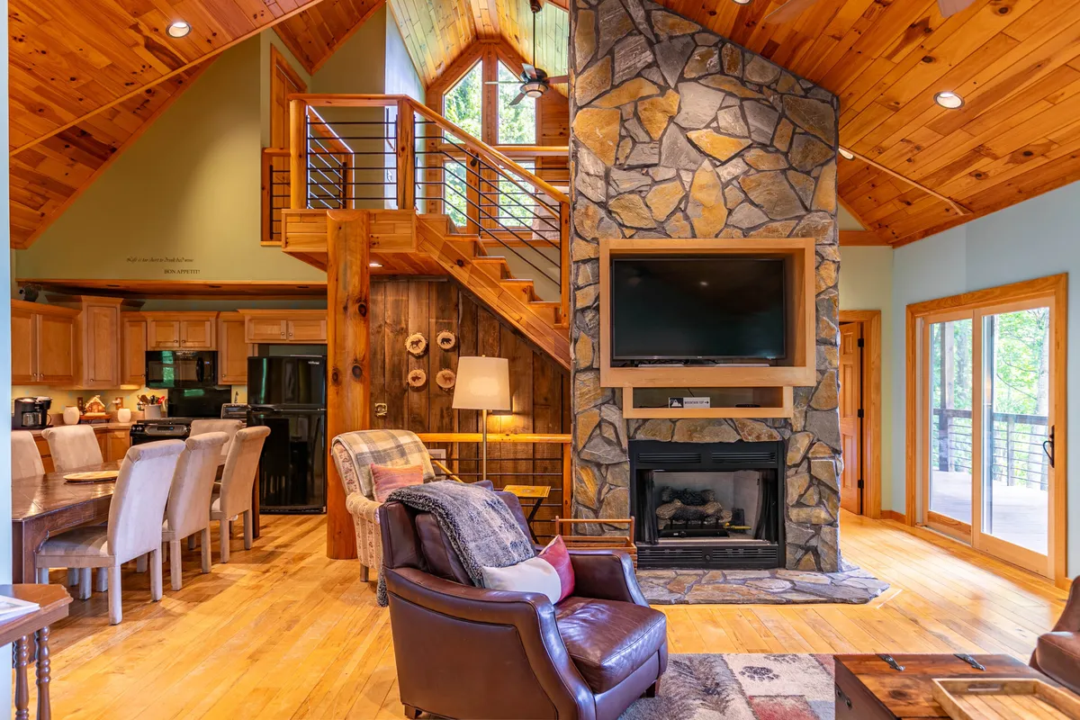 The scene features a rustic stone fireplace, exposed wooden beams, a large dining table, and a comfortable leather armchair. The space is designed for gathering, whether for meals, games, or cozy nights by the fire.