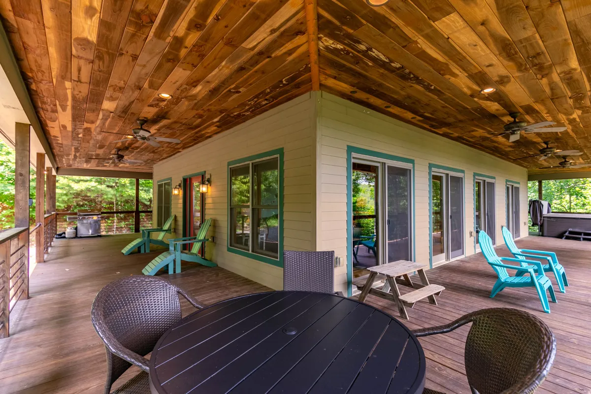 A generous wooden deck with a set of adirondack chairs and a large outdoor dining table sits beneath a wooden ceiling, framed by big windows and surrounded by trees. The arrangement suggests an inviting space for al fresco dining, relaxation, and group gatherings, with sunset or stargazing opportunities.