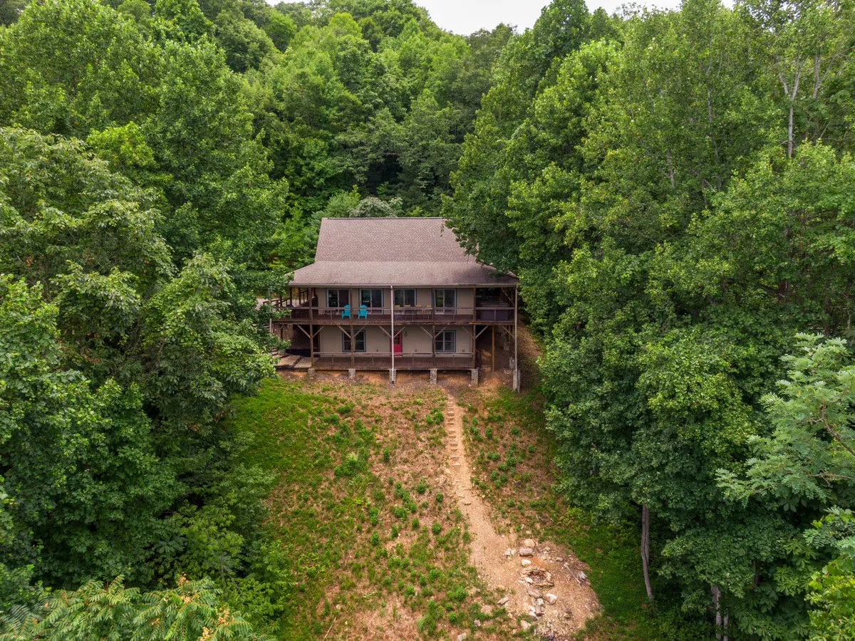 The image features a two-story cabin embraced by tall trees, with a pathway leading to the house—an ideal setting for outdoor gatherings. The natural surroundings evoke a sense of seclusion and the opportunity for bonfires or yard activities, hinting at the fire pit and wooded yard mentioned in the text.